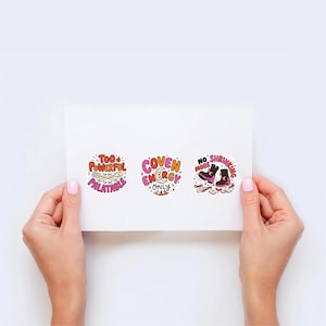 Op de afbeelding: Drie ronde stickers op een wit vel. De eerste bevat de tekst 'TOO POWERFUL TO BE PALATABLE' met een vork en lepel. De tweede zegt 'COVEN ENERGY ONLY' met een maansikkel. De derde toont 'NO MORE SHRINKING' met twee zwarte laarzen.