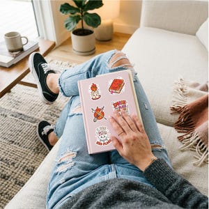 Op de afbeelding: Een lichtroze notitieboek met vijf verschillende stickers: een kaars met een vlam, een rood boek met een gezicht, een bezem gekruist met een zwaard, de tekst SMASH THE GLASS CEILING en een kristallen bol met de tekst I SEE RED FLAGS.