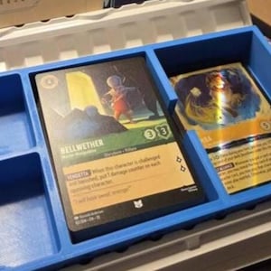 May include: A blue plastic organizer with compartments holding illustrated game cards. One card features a figure in a landscape, labeled "Bellwether." The other card has a golden, shimmering design. The organizer is inside a larger white container.