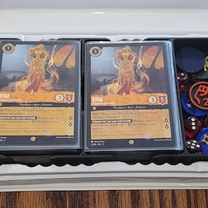 May include: A game set with two trading cards featuring a golden-haired figure, dice, and tokens. The cards have text and a gold and orange color scheme. The set is in a black tray within a white box, on a wooden surface.