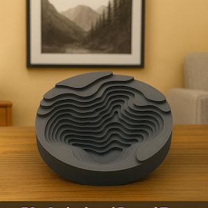May include: A grey, layered, circular decorative object resembling a topographical map. The object has a series of concentric, curved layers, creating a three-dimensional effect. The piece is made of a solid, matte material and sits on a wooden surface.