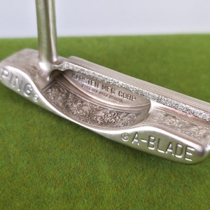 May include: A silver-colored golf putter with the text "PING" and "A-BLADE" visible. The putter has intricate floral engravings and the text "KARSTEN MFG CORP". The putter is on a green surface.