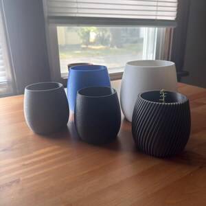 May include: Five small, textured plant pots in shades of dark grey, blue, black, white, and a black spiral design. One pot contains a small green plant. The pots are displayed on a wooden surface.