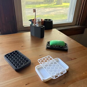 May include: A collection of 3D-printed bathroom accessories, including a black soap dish with a green bar of soap, a toothbrush holder, and a white and black container with a honeycomb pattern. The items are arranged on a wooden table.