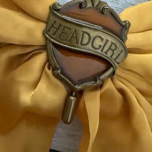 May include: A mustard yellow fabric bow with a decorative bronze-colored shield-shaped pin. The pin has the word "HEADGIRL" in a banner across the top. The pin has a brown center and a bronze-colored pin backing.