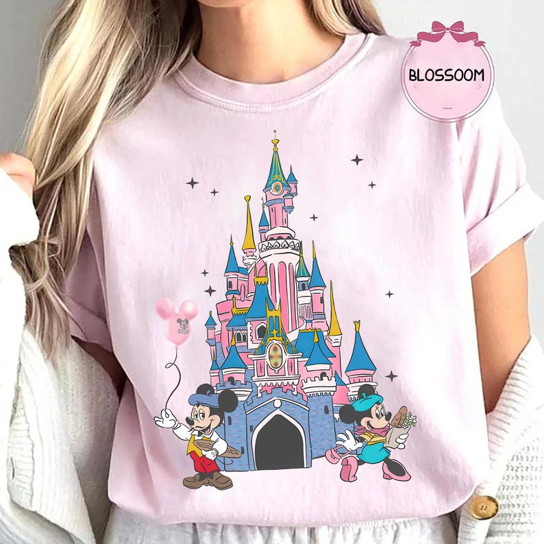 Może przedstawiać: Jasnor&oacute;żowa koszulka z ilustracją zamku z bajki, Myszki Miki i Myszki Minnie. Myszka Miki trzyma balon, a Myszka Minnie niesie kwiaty. Koszulka ma napis "BLOSSOM" w r&oacute;żowym okręgu.