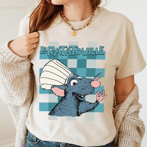 Könnte beinhalten: Beiges T-Shirt mit einer Cartoon-Illustration einer Ratte mit Kochmütze und dem Text "LA RATATOUILLE" darüber. Die Ratte ist vor einem blau-weiß karierten Hintergrund platziert.
