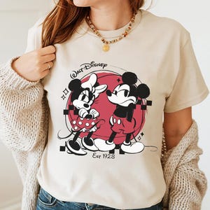 May include: Beige t-shirt featuring a black and red graphic of Mickey and Minnie Mouse. The design includes the text "Walt Disney" and "Est 1928". Minnie is wearing a polka dot dress, and Mickey is wearing red shorts.