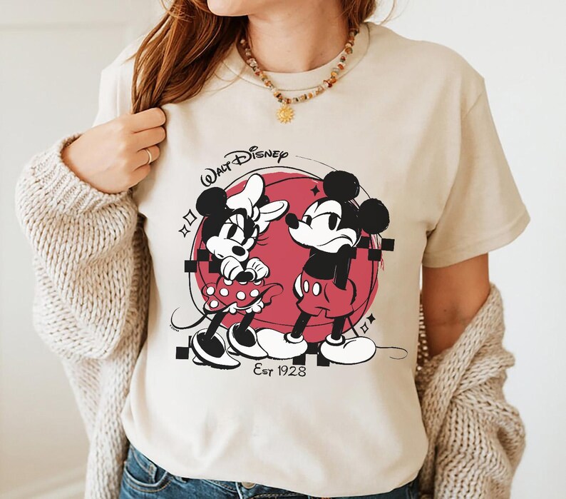 May include: Beige t-shirt featuring a black and red graphic of Mickey and Minnie Mouse. The design includes the text "Walt Disney" and "Est 1928". Minnie is wearing a polka dot dress, and Mickey is wearing red shorts.