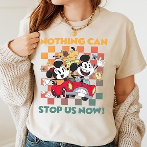 May include: Beige t-shirt featuring a colorful graphic of Mickey and Minnie Mouse, Pluto, and other cartoon characters in a red car. The text "NOTHING CAN STOP US NOW!" is printed above and below the graphic.