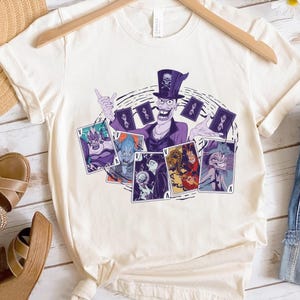 May include: Cream-colored t-shirt featuring a cartoon illustration of a villain character in a purple suit and top hat, holding playing cards with other villain characters. The shirt is hanging on a wooden hanger.