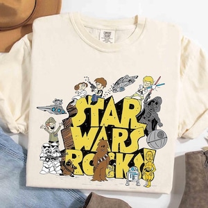 Puede incluir: Camiseta color crema con un gráfico de dibujos animados de personajes de Star Wars. Las palabras "STAR WARS ROCKS" están en letras amarillas. El diseño incluye personajes como Darth Vader, Chewbacca y C-3PO.