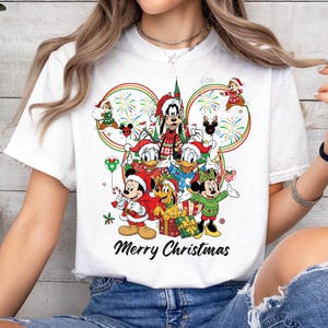 May include: White t-shirt featuring a festive Disney design with Mickey Mouse, Minnie Mouse, Donald Duck, Goofy, and other characters in holiday attire. The words "Merry Christmas" are printed below the characters.