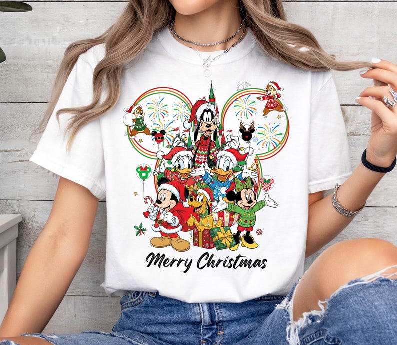 May include: White t-shirt featuring a festive Disney design with Mickey Mouse, Minnie Mouse, Donald Duck, Goofy, and other characters in holiday attire. The words "Merry Christmas" are printed below the characters.