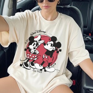 May include: Cream-colored t-shirt featuring a graphic of Mickey and Minnie Mouse in front of a red circle. The Walt Disney logo is above the graphic, with "Est 1928" below. The shirt has short sleeves.