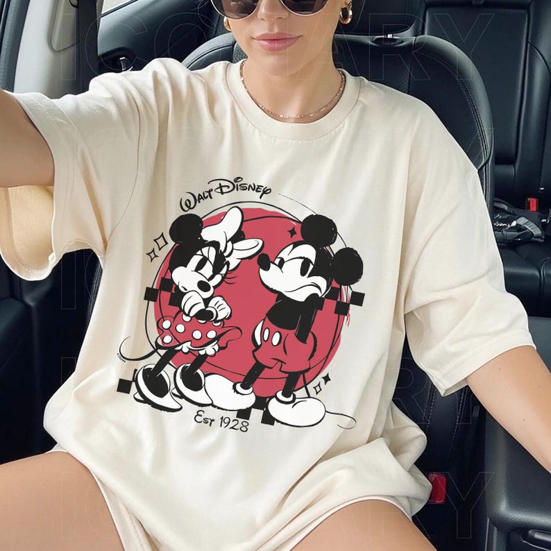 May include: Cream-colored t-shirt featuring a graphic of Mickey and Minnie Mouse in front of a red circle. The Walt Disney logo is above the graphic, with "Est 1928" below. The shirt has short sleeves.