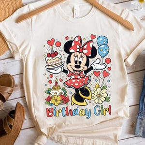 Peut inclure: T-shirt cr&egrave;me avec un dessin d'une souris de dessin anim&eacute; en robe &agrave; pois rouges, tenant un g&acirc;teau et des ballons. Le texte "Birthday Girl" est imprim&eacute; en lettres color&eacute;es en dessous. Le t-shirt est suspendu &agrave; un cintre en bois.