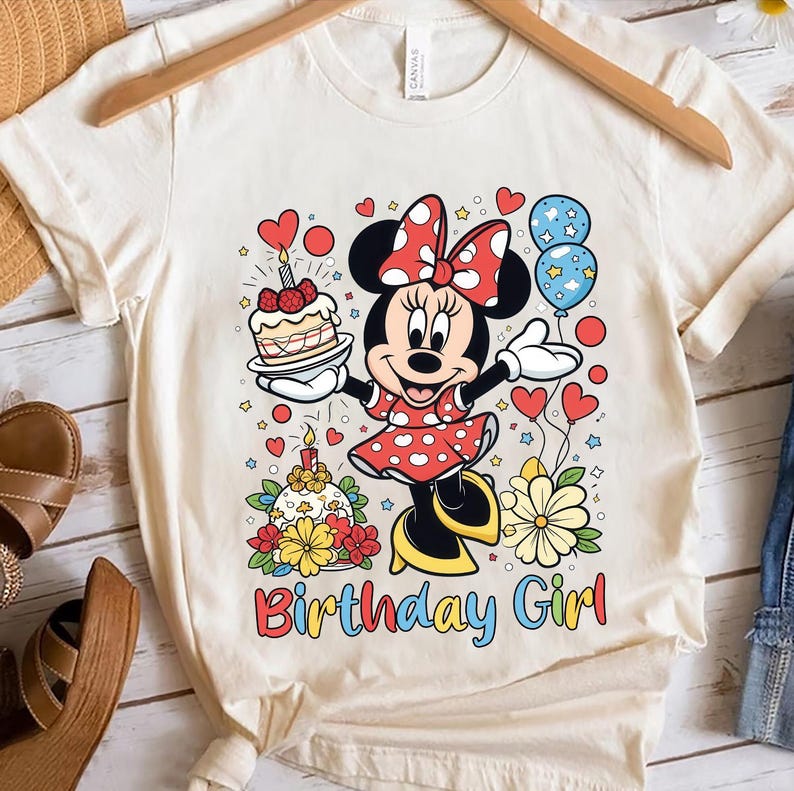 Peut inclure: T-shirt cr&egrave;me avec un dessin d'une souris de dessin anim&eacute; en robe &agrave; pois rouges, tenant un g&acirc;teau et des ballons. Le texte "Birthday Girl" est imprim&eacute; en lettres color&eacute;es en dessous. Le t-shirt est suspendu &agrave; un cintre en bois.