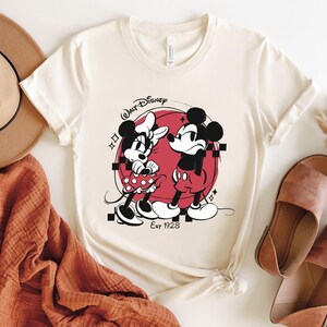 May include: Cream-colored t-shirt featuring a graphic of Mickey and Minnie Mouse in black and red, set against a red circle. The shirt includes the text "Walt Disney" and "Est 1928".