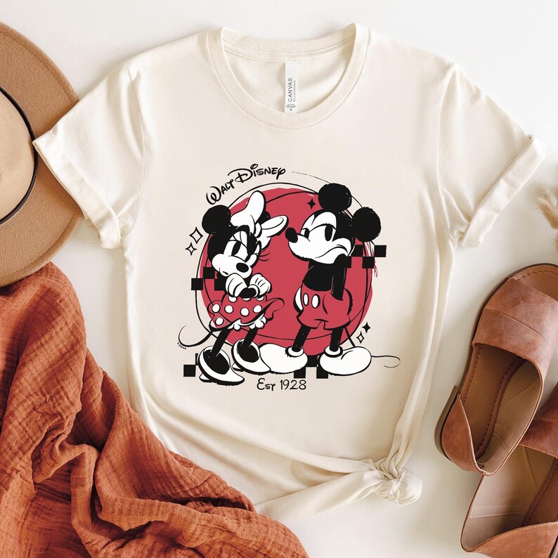 May include: Cream-colored t-shirt featuring a graphic of Mickey and Minnie Mouse in black and red, set against a red circle. The shirt includes the text "Walt Disney" and "Est 1928".