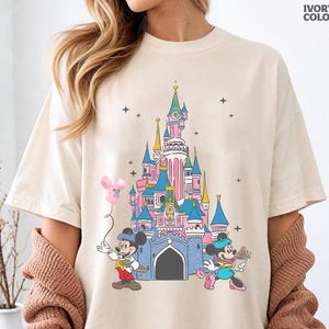 Może przedstawiać: Kremowa koszulka z kolorową grafiką zamku Disneya z Myszka Miki i Myszka Minnie. Miki trzyma r&oacute;żowy balon, a Myszka Minnie niesie kwiaty. Tekst "IVORY COLOR" jest wydrukowany w prawym g&oacute;rnym rogu.