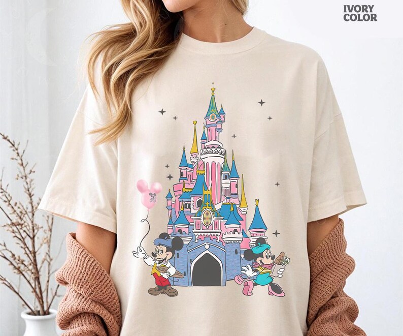 Może przedstawiać: Kremowa koszulka z kolorową grafiką zamku Disneya z Myszka Miki i Myszka Minnie. Miki trzyma r&oacute;żowy balon, a Myszka Minnie niesie kwiaty. Tekst "IVORY COLOR" jest wydrukowany w prawym g&oacute;rnym rogu.