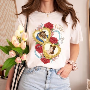 Puede incluir: Camiseta blanca con un diseño colorido de La Bella y la Bestia de Disney. El diseño presenta a Bella y la Bestia en marcos en forma de corazón, rodeados de rosas rojas y el título de la película.