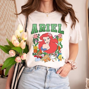 Può includere: T-shirt bianca con la scritta "ARIEL" in lettere verdi a blocchi sopra una grafica colorata di Ariel de La Sirenetta. La grafica include Ariel, fiori e creature marine. La maglietta è abbinata a jeans blu.