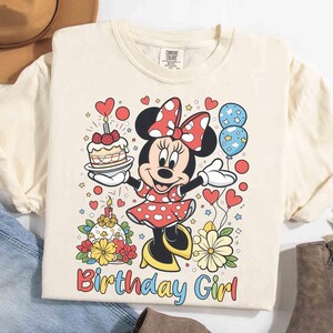 Peut inclure: T-shirt cr&egrave;me avec un motif Minnie Mouse color&eacute;. Minnie tient un g&acirc;teau et des ballons, avec les mots "Birthday Girl" en dessous. Le motif comprend des c&oelig;urs, des fleurs et un g&acirc;teau d'anniversaire.