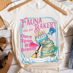 May include: Cream-colored t-shirt featuring a whimsical "Fauna Bakery" design. The graphic includes a layered cake with candles, a broom, and the text "Once Upon A Dream" and "Baked With Love & Magic".