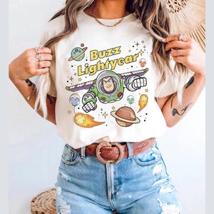 Puede incluir: Camiseta color crema con un gráfico colorido de Buzz Lightyear en pose de vuelo, con las palabras "Buzz Lightyear" arriba. El diseño incluye planetas, estrellas y otros elementos temáticos del espacio. La camiseta se combina con pantalones cortos de mezclilla azules y un cinturón marrón.