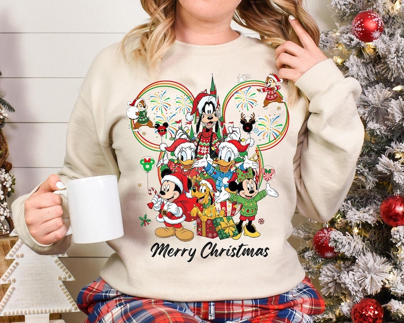 May include: A cream-colored sweatshirt featuring a festive Disney Christmas design. The graphic includes Mickey, Minnie, Donald, Goofy, and other characters in Santa hats, with the words "Merry Christmas" below.