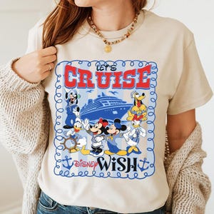 Könnte beinhalten: Beiges T-Shirt mit einem Aufdruck "Let's Cruise Disney Wish". Das Design zeigt ein blaues Kreuzfahrtschiff und Zeichentrickfiguren wie Mickey Mouse, Minnie Mouse, Donald Duck und Pluto. Die Grafik ist von einem blauen Rahmen mit Ankern umgeben.