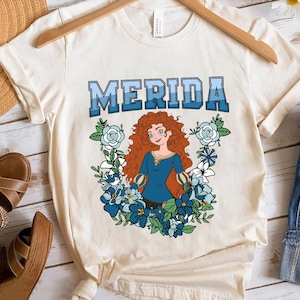 Könnte beinhalten: Cremefarbenes T-Shirt mit dem Namen "MERIDA" in blauen Blockbuchstaben. Darunter eine Cartoon-Illustration einer Frau mit roten Locken, die ein blaues Kleid trägt, umgeben von blauen und weißen Blumen.