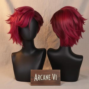 May include: Two red-burgundy wigs with short, layered styles are displayed on black mannequin heads. The wigs are positioned side-by-side, showcasing the front and back views. A wooden sign with the text "Arcane VI" is placed in front of the mannequins.