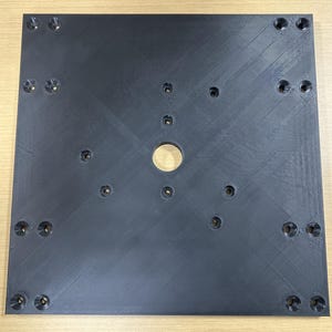 Puede incluir: Una placa cuadrada negra con múltiples agujeros y una abertura circular central. La placa tiene una superficie texturizada con líneas diagonales. Hay varios agujeros más pequeños alrededor del perímetro y el centro.