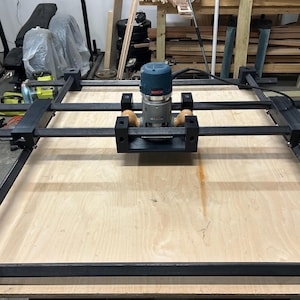 Puede incluir: Una máquina CNC para carpintería con una fresadora Bosch. La máquina tiene un marco de metal negro y se asienta sobre una superficie de madera grande y de color claro. La fresadora es azul y plateada.