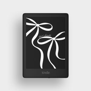 Puede incluir: Un lector de libros electrónicos Kindle negro con una pantalla negra que muestra un diseño de lazo blanco y el texto "blair's kindle". La palabra "kindle" está impresa en la parte inferior del dispositivo.