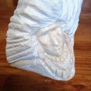 May include: A carved stone sculpture of a Native American chief. The sculpture is off-white with detailed carving of the face, headdress, and neck adornment. The sculpture is set against a wooden background.
