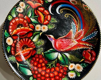 May include: A hand-painted decorative plate with a black background. The plate features a vibrant design of a red bird with a long, colorful tail, surrounded by red flowers, white daisies, and red berries. The rim is accented with gold dots.