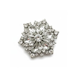Puede incluir: Un broche plateado con diseño floral. El broche presenta un grupo central de cristales grandes y transparentes rodeados de piedras más pequeñas y brillantes, creando un deslumbrante efecto de estrella. El broche está sobre un fondo blanco.
