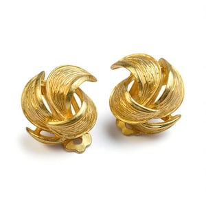 Puede incluir: Un par de pendientes dorados de clip. Cada pendiente presenta un diseño arremolinado y texturizado con elementos curvos en forma de hoja. Los pendientes tienen un acabado brillante y pulido y una estética vintage clásica.