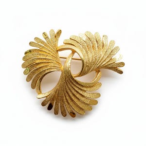 May include: A gold-toned brooch with a textured, swirling design. The brooch features three fan-like elements that curve inward, creating a dynamic, eye-catching piece. The pin is a classic accessory.