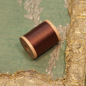 May include: A spool of brown thread wound around a wooden bobbin. The bobbin is sitting on a green fabric with gold trim.
