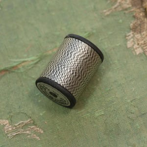 May include: A spool of silver and black thread with a black rim. The thread is tightly wound, and the spool is resting on a green fabric surface. The spool has text on the side.