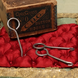 May include: An antique box labeled "ONE GROSS NO 0 C.B.G. BUTTON HOOKS" sits on a red, tufted cushion. Two metal button hooks are placed on the cushion, with one inserted. The box is brown with black and white text.