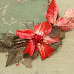 May include: A close-up of a handmade floral arrangement. The arrangement features a vibrant red flower with a textured surface, surrounded by gray and pink leaves. The background is a muted green with a decorative pattern.