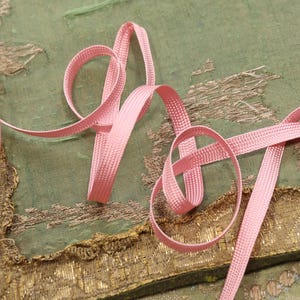 May include: Pink satin ribbon with a textured surface. The ribbon is folded and twisted into a decorative shape.