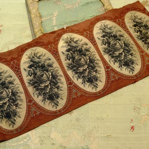 May include: A rectangular strip of fabric featuring a repeating pattern of four cream-colored ovals. Each oval contains a detailed black floral bouquet with roses. The background of the strip is a deep rust-orange with a dotted border.