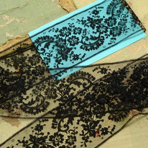 May include: A length of black lace trim featuring a detailed floral pattern with scalloped edges. The delicate mesh fabric is displayed partially over a bright blue rectangular card, showcasing the intricate needlework and sheer texture of the material.